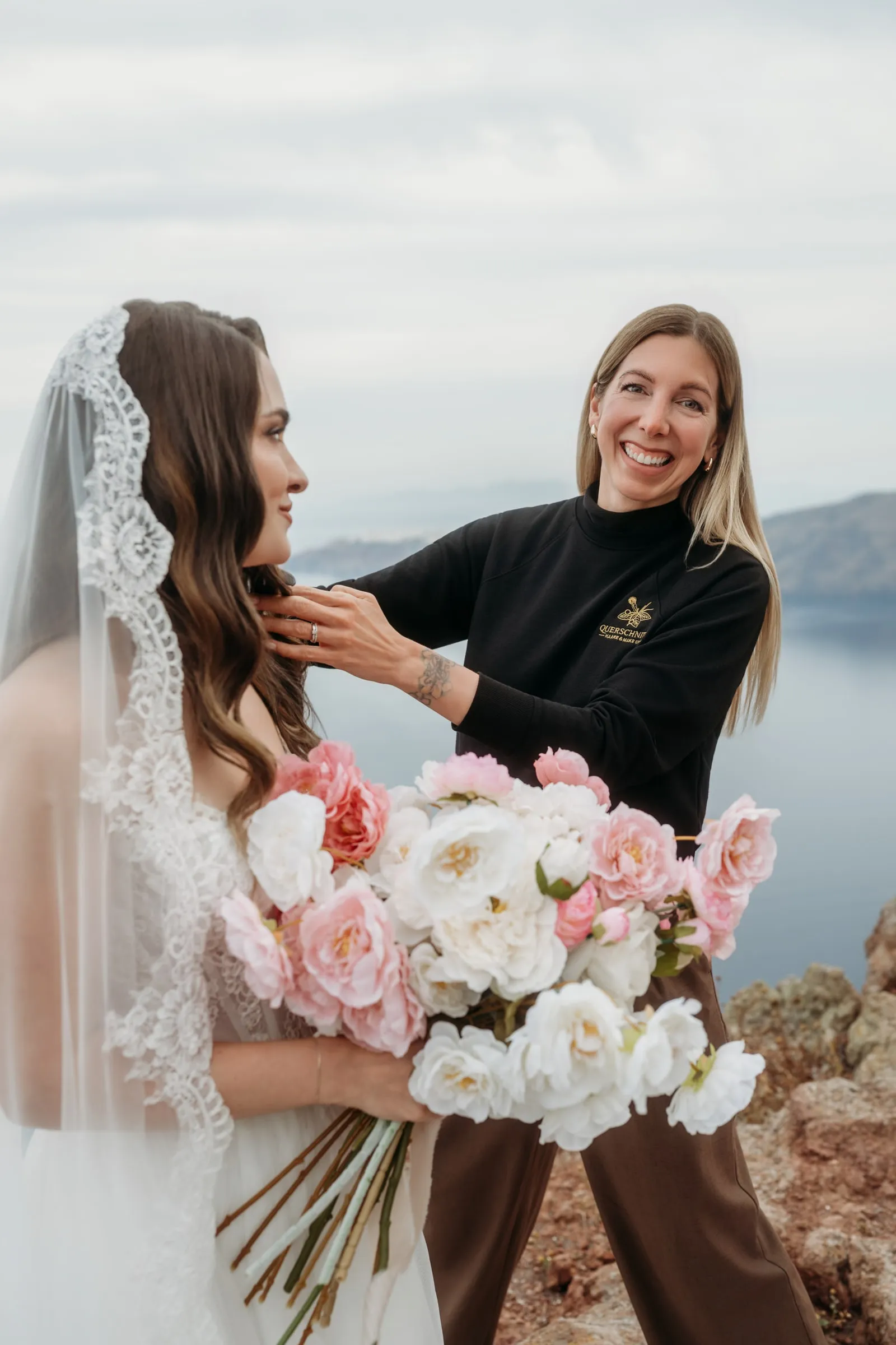 Friseurmeisterin Jasmin Werner steckt einer lachenden Braut mit Schleier und Brautstrauß auf Santorin die Haare