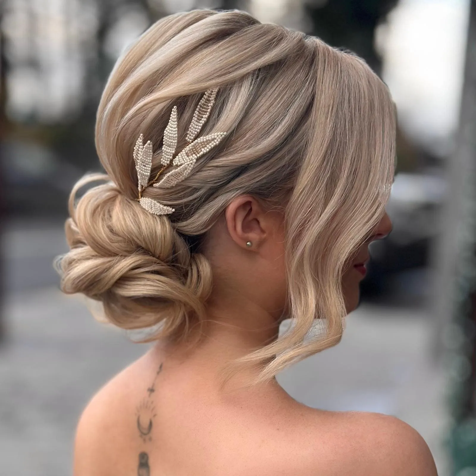 Bridal Hair von Kasia Fortuna: seitlicher Low Bun in Blond mit glattem Volumen und Haarschmuck aus Perlenblättern